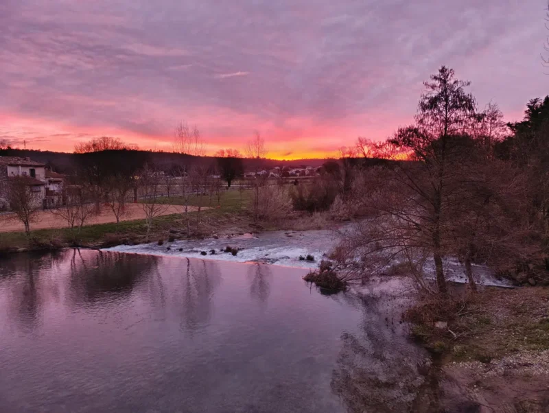 Photo d'un levé de soleil sur un petit fleuve coupé d'une petite cascade artificielle, avec des arbres sans feuilles, des maisons en arrière-plan et un ciel aux tons roses et orangés se reflétant dans l'eau.