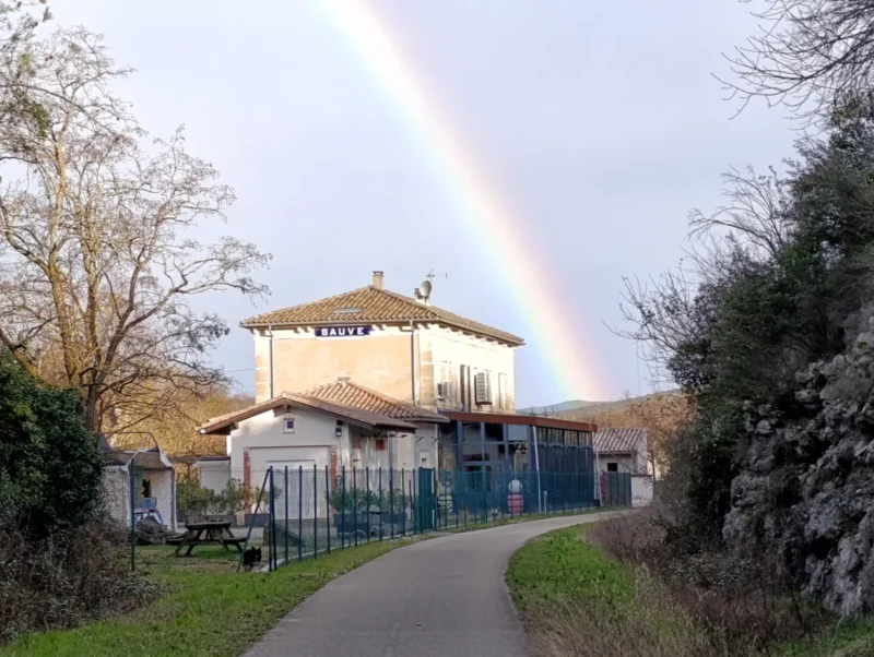 Photo prise depuis une voie verte. Sur la gauche de la piste cyclable, une ancienne gare derrière un grillage sur laquelle apparaît le nom de « Sauve ». Un bel arc-en-ciel vient frôler le bâtiment.