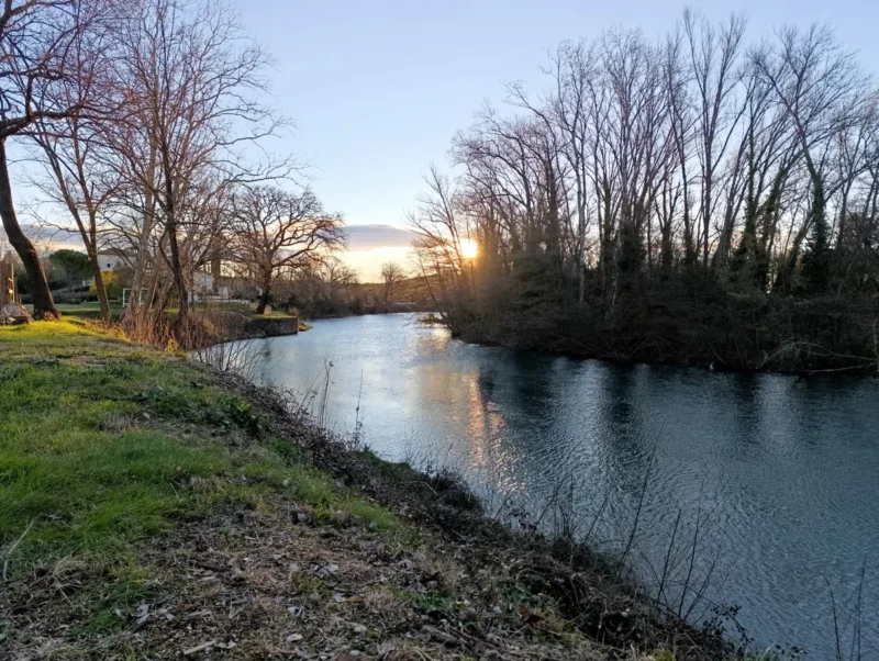 Photo d'un cours d'eau calme entouré d'arbres sans feuilles, avec un coucher de soleil visible à travers les branches, reflétant une lumière dorée sur la surface de l'eau et une rive herbeuse en premier plan. On distingue quelques habitations sur la gauche de l'image.