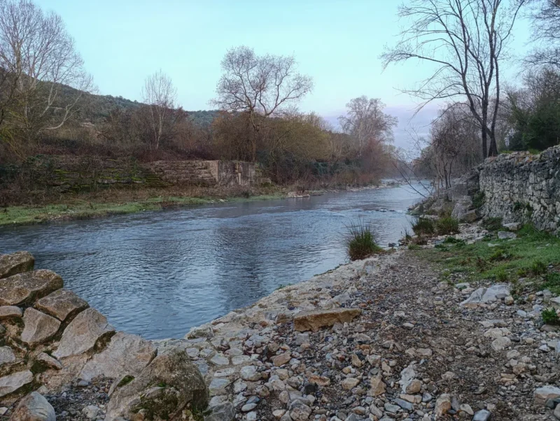 Photo d'un petit fleuve dont le niveau est bas, entouré d’arbres et de végétation, avec un ancien mur au premier plan et des vestiges en pierres sur les deux rives. Le ciel est dégagé dans une faible lumière matinale.