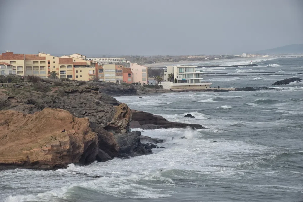 Photo de falaises volcaniques sous l'assaut des vagues. Plus loin, des immeubles bordent la côte d'une Méditerranée agitée.
