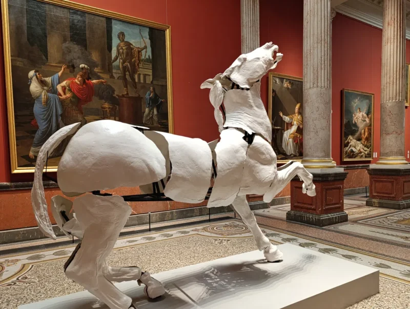 Photo de la statue contemporaine d'un grand cheval blanc dans une salle de musée, contrastant avec d'anciennes peintures et des colonnes en arrière-plan.