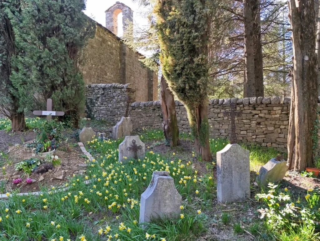 Photo de quelques tombes dans un parterre de fleurs. En arrière-plan, une ancienne chapelle et quelques arbres.