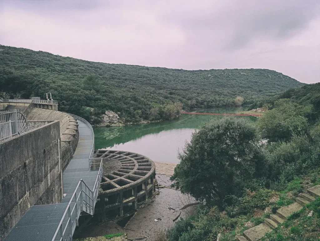 Photo légèrement filtré. Au premier plan, le déversoir d'un barrage et sa retenue d'eau, couleur verdâtre, bordée d'arbres. Le ciel est chargé de nuages.