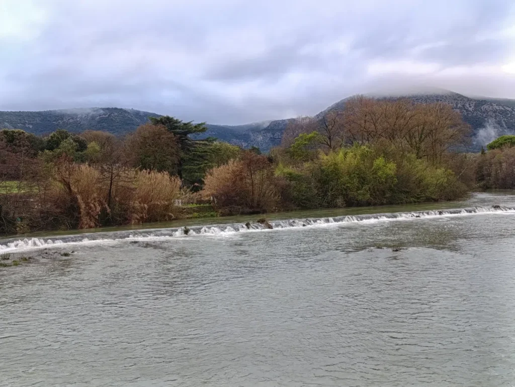 Photo du Vidourle prise d'un pont qui le surplombe. Une petite cascade et une rangée d'arbres et de végétation aux teintes automnales en second plan. En arrière-plan, un petit massif sous les nuages.