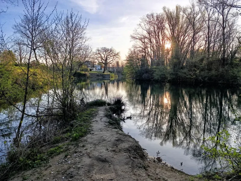 Sur la rive d'un petit fleuve côtier. En face, sur la même rive, on perçoit une maison au loin. Sur la rive opposée, quelques arbres derrière lesquels pointe le soleil. Une petite sente s'avance vers l'eau sur une petite pointe de terre. Le ciel est dégagé avec un léger voile nuageux.