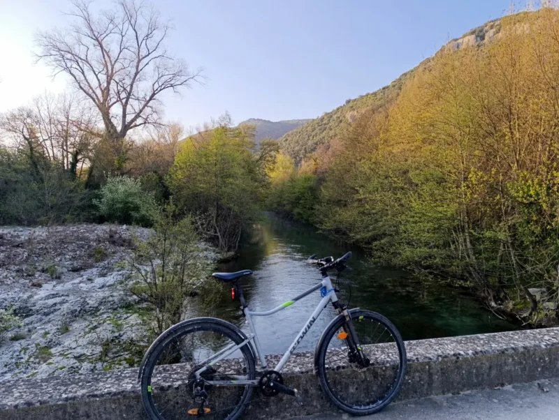 Photo d'un vélo qui repose sur le petit parapet d'un petit pont qui enjambe un fleuve aux rives arborées. Sur la gauche, une dalle de pierre et un arbre sans feuille. Le ciel est bleu et dégagé dans une lumière matinale.