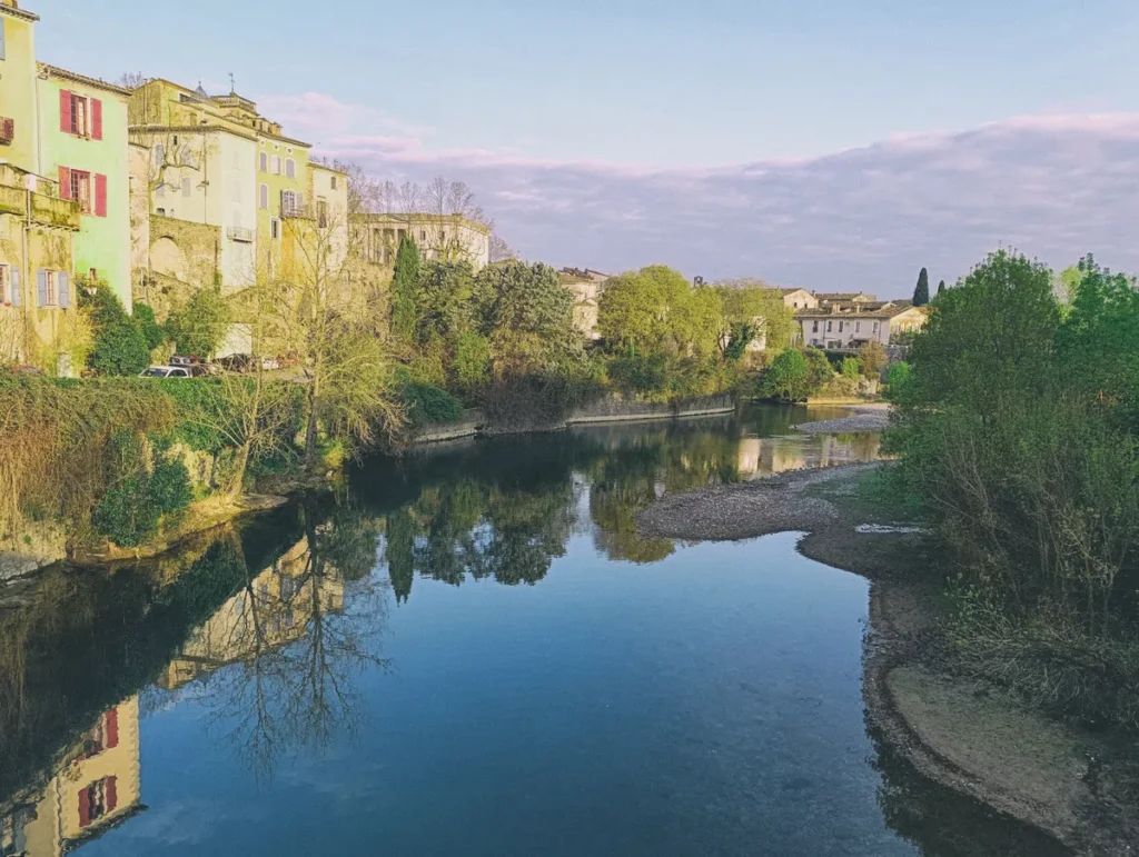 Un petit village sur les rives d'un petit fleuve. Des maisons sont visibles sur la gauche de l'image, dont l'image se reflète en partie sur le cours d'eau. Le ciel est bleu avec un banc de nuages au loin.