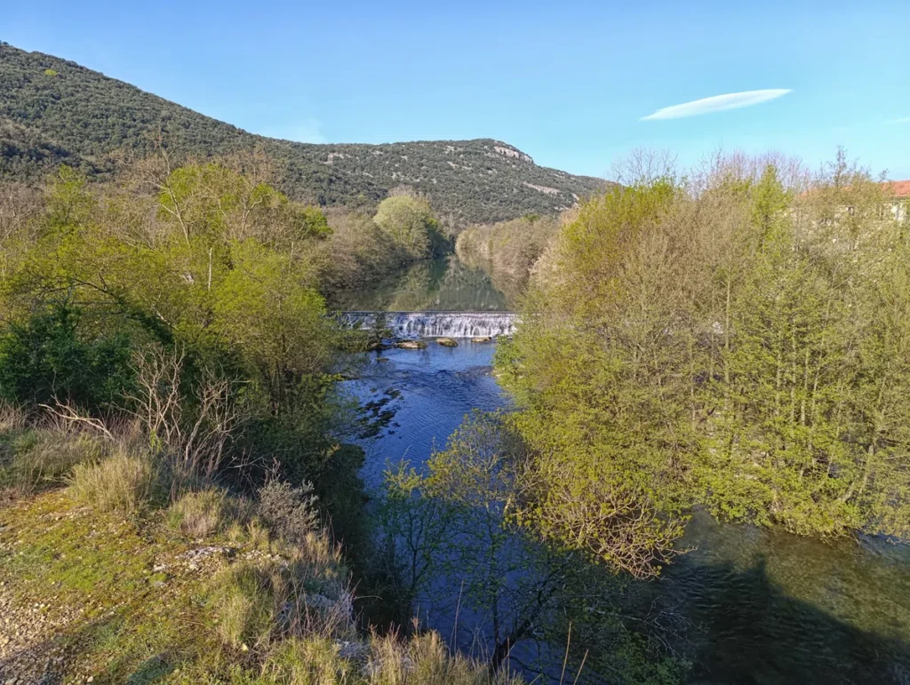 Une cascade sur un petit fleuve visible entre les arbres. Plus loin, un petit massif. Le ciel au-dessus est bien bleu.