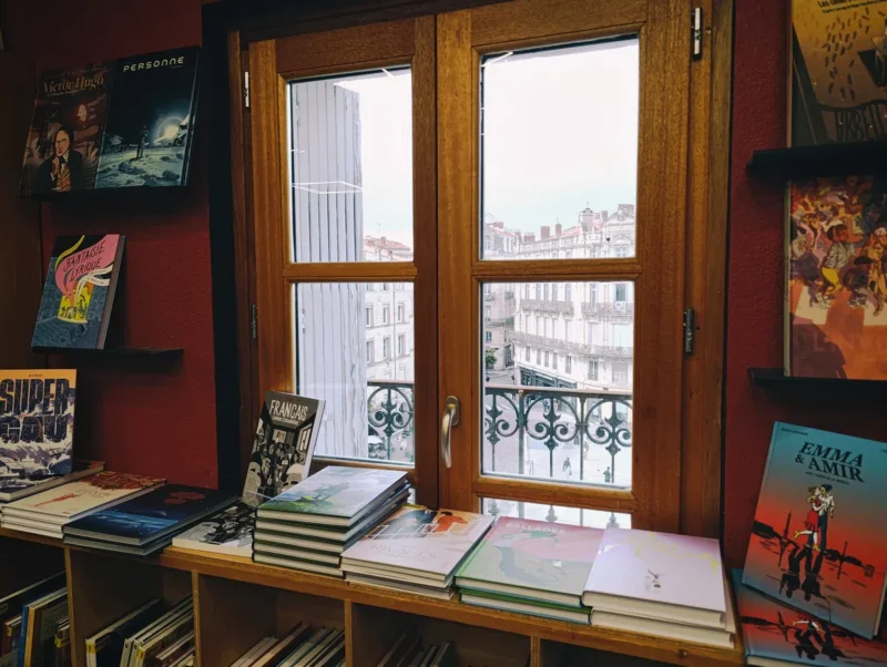 Photo dans une librairie. Des livres posés sur une table devant une fenêtre, dont on perçoit la vue sur la ville et ses immeubles.
