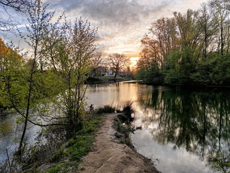 Sur la rive d'un petit fleuve côtier. En face, sur la même rive, on perçoit une maison au loin. Sur la rive opposée, quelques arbres derrière lesquels pointe le soleil. Une petite sente s'avance vers l'eau sur une petite pointe de terre. Le ciel est dégagé avec un léger voile nuageux.