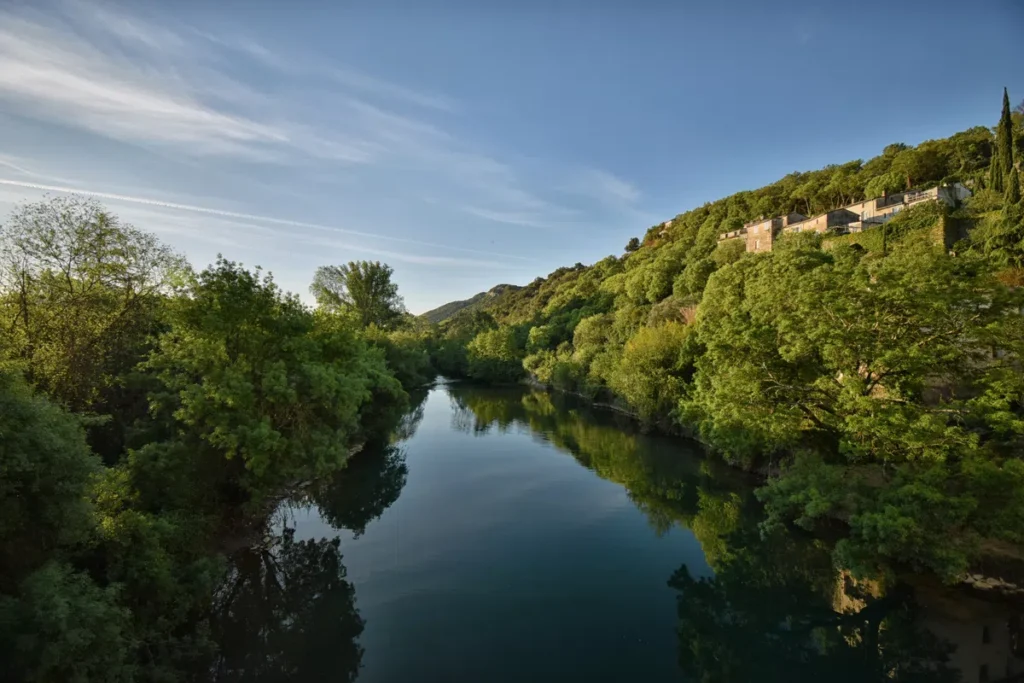 Photo d'un fleuve paisible aux rives arborées sous un ciel bleu. On distingue une bâtisse sur la droite, en hauteur sur un petit massif.