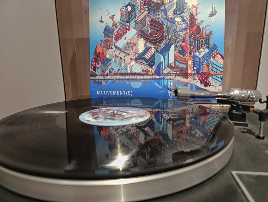 Photo d'une œuvre exposée dans une galerie d'art. Un tourne-disque en fonctionnement au premier plan, avec un disque vinyle noir et le bras de lecture posé dessus. La surface du disque reflète l’image derrière. En arrière-plan, une pochette illustrée montre une ville stylisée et colorée avec bâtiments et structures mécaniques, sur fond bleu, avec le mot « MOUVEMENT(S) » visible en bas.